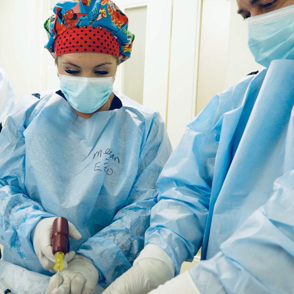 Two medical professionals in blue scrubs and masks are practicing a procedure, with one holding a device while the other assists, in a clinical setting.